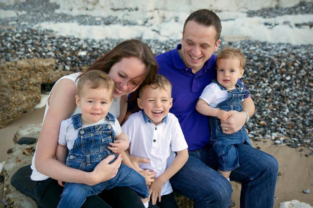Eastbourne_family_photography_beach_powell-ful_photography.4 mum and dad sitting on the pebbly beach with there three young boys on there laps smiling and laughing
