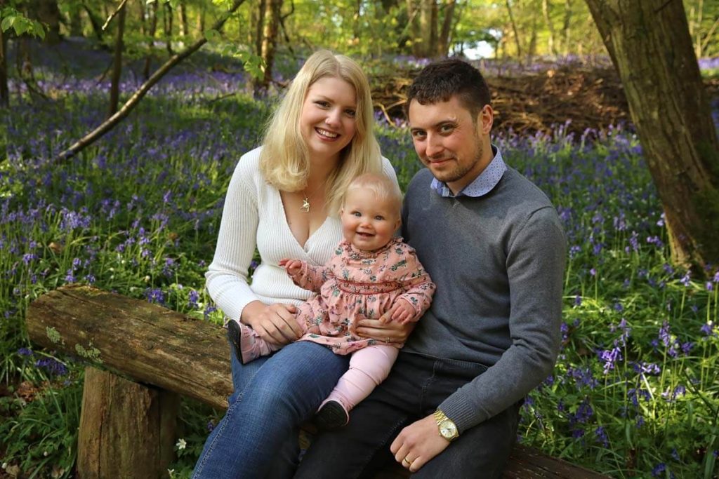 Eastbourne_family_photographer_bluebells_powell-ful_photography.5 mum and dad sitting on bench in bluebell woods hold there little girl