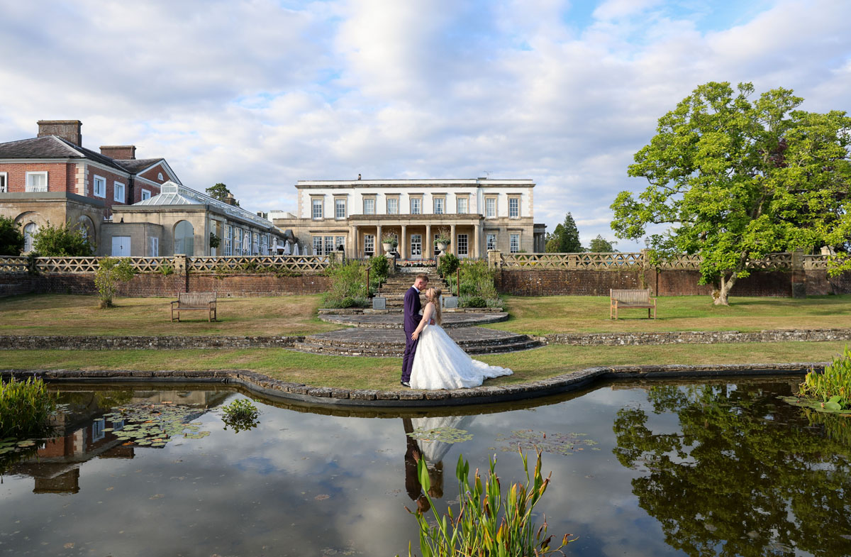 bride and groom standing in front of a pond, Buxted Park Behind them